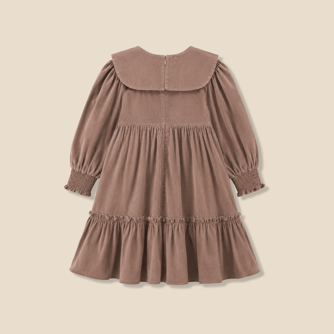 A young girl with her hair in a bun wears a brown corduroy dress with a large ruffled collar and bow, standing against a solid beige background.