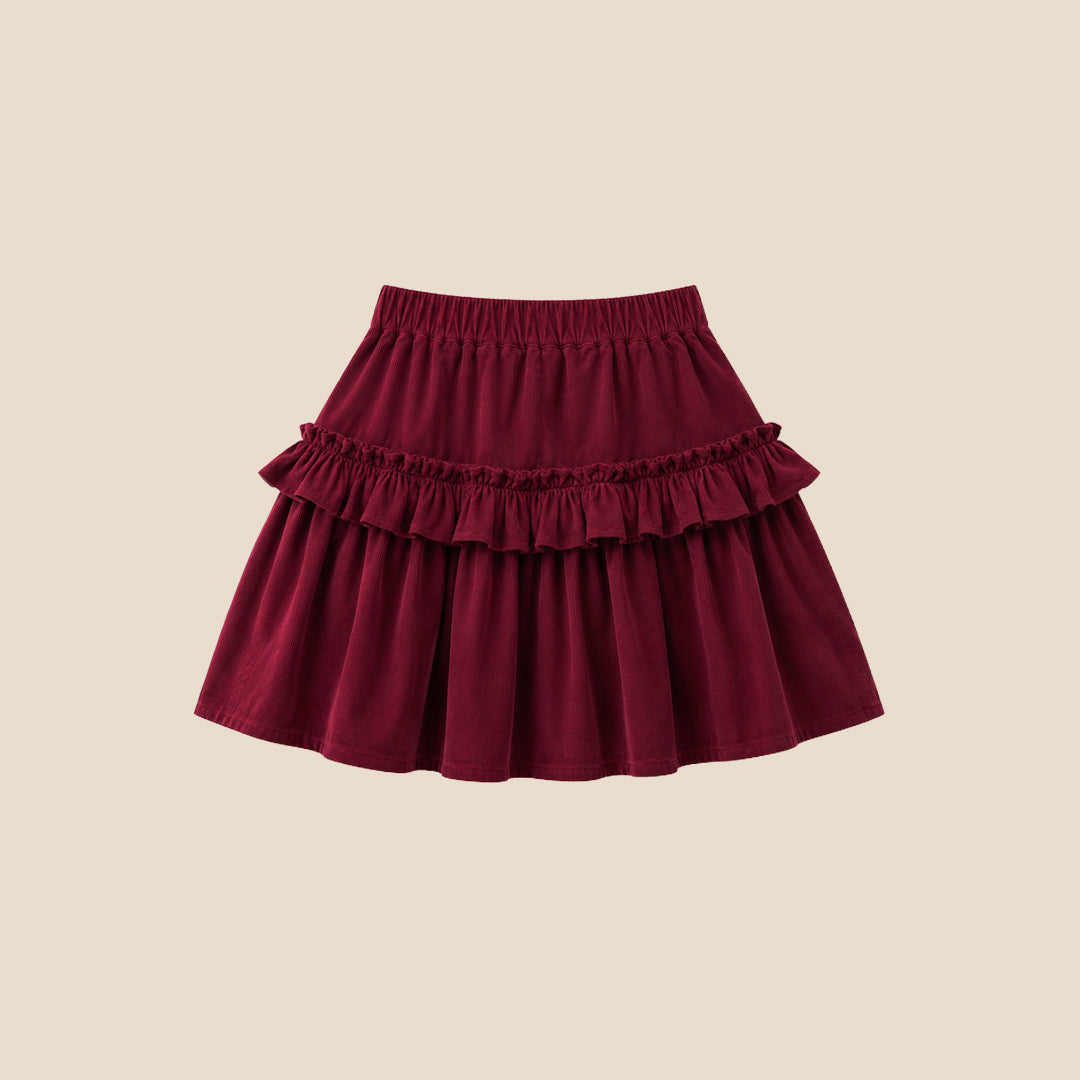 A child wearing a tiered burgundy skirt with ruffles and a white puff-sleeve top featuring a pink plaid bow graphic, photographed against a plain beige background.
