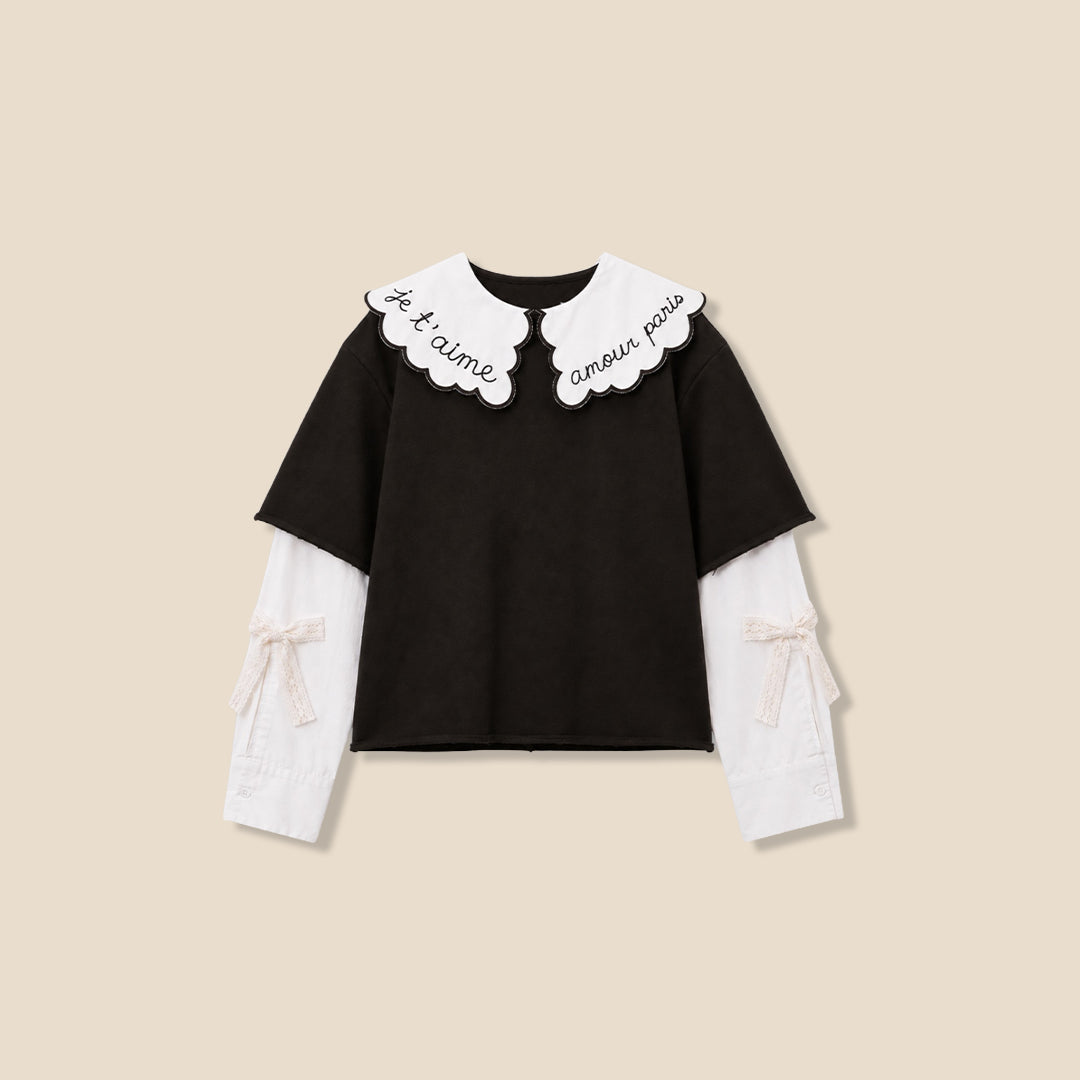 A young girl with wavy light brown hair is wearing a dark brown short-sleeve top with a large white ruffled collar featuring black cursive embroidery, layered over a white long-sleeve shirt, paired with brown and black leopard-print pants, against a solid beige background.