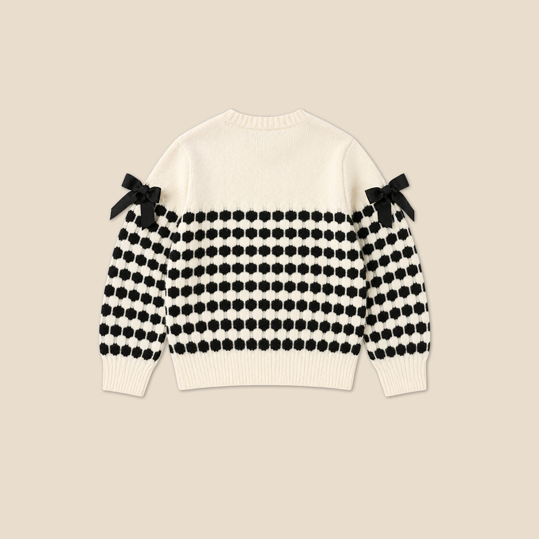 A young girl with her hair in a bun wears a cream knit cardigan with black polka-dot patterned sleeves and black ribbon bows on the shoulders, paired with a dark blue ruffled skirt, against a solid beige background.