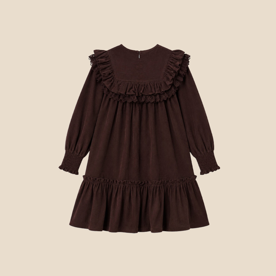 A young girl with her hair in a high bun models a dark brown long-sleeve dress with ruffled details, set against a solid light beige background.