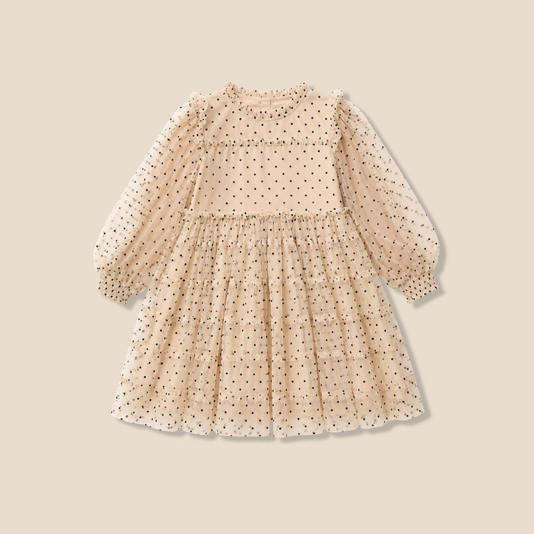 A young girl with her hair in a bun wears a cream-colored, long-sleeved tiered dress with black polka dots, standing against a solid beige background.