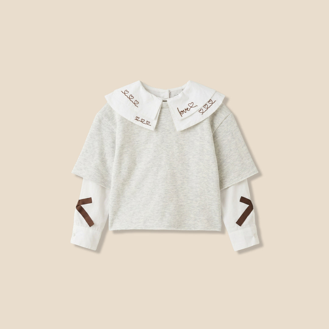 A young girl with her hair in a ponytail models a light gray long-sleeve top featuring a large white collar with brown embroidered details and matching brown accents on the sleeves, paired with blue jeans against a solid beige background.