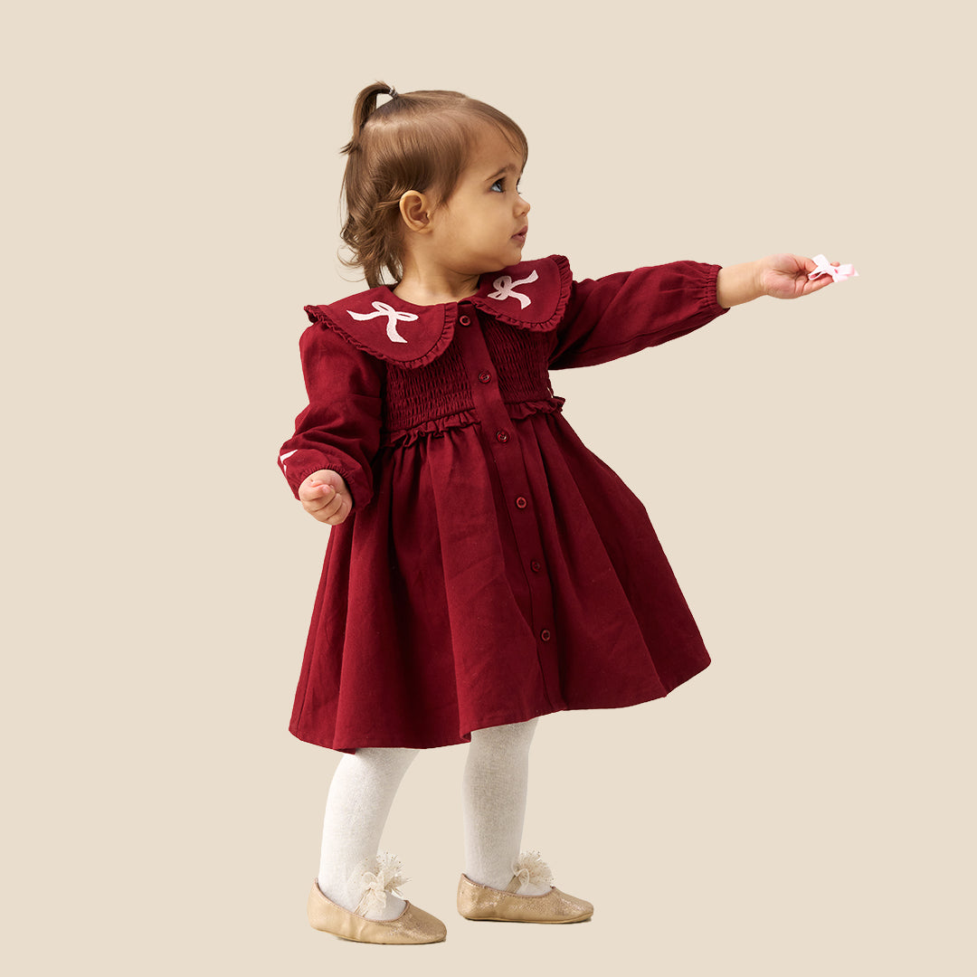 A toddler girl wearing a burgundy corduroy dress with a large white ruffled collar decorated with bows, white tights, and beige ballet flats, standing against a solid beige background.