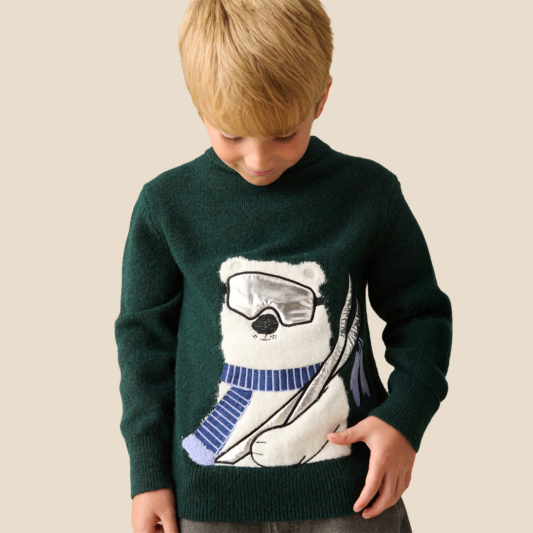 A young boy with blond hair wearing a dark green knit sweater featuring a large white polar bear graphic with ski goggles and a blue striped scarf, set against a solid beige background.