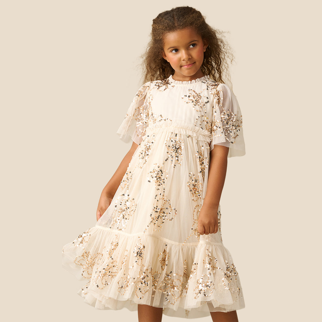 A young girl with curly brown hair wearing a tiered cream dress decorated with gold sequins and floral embroidery, standing against a solid beige background.