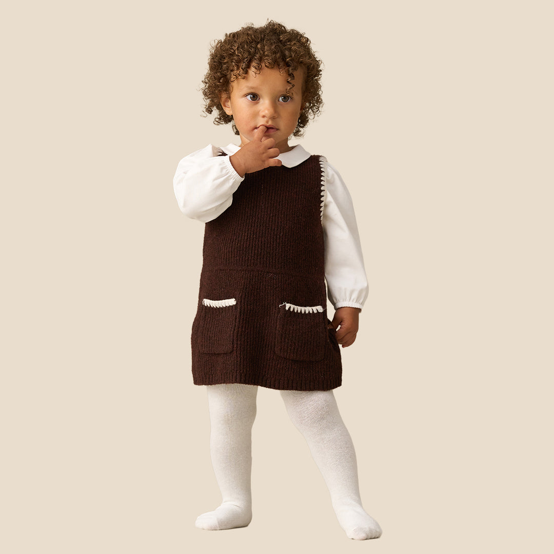 A toddler with curly brown hair wearing a dark brown corduroy pinafore dress over a white long-sleeve collared shirt and white tights, standing against a solid light beige background.