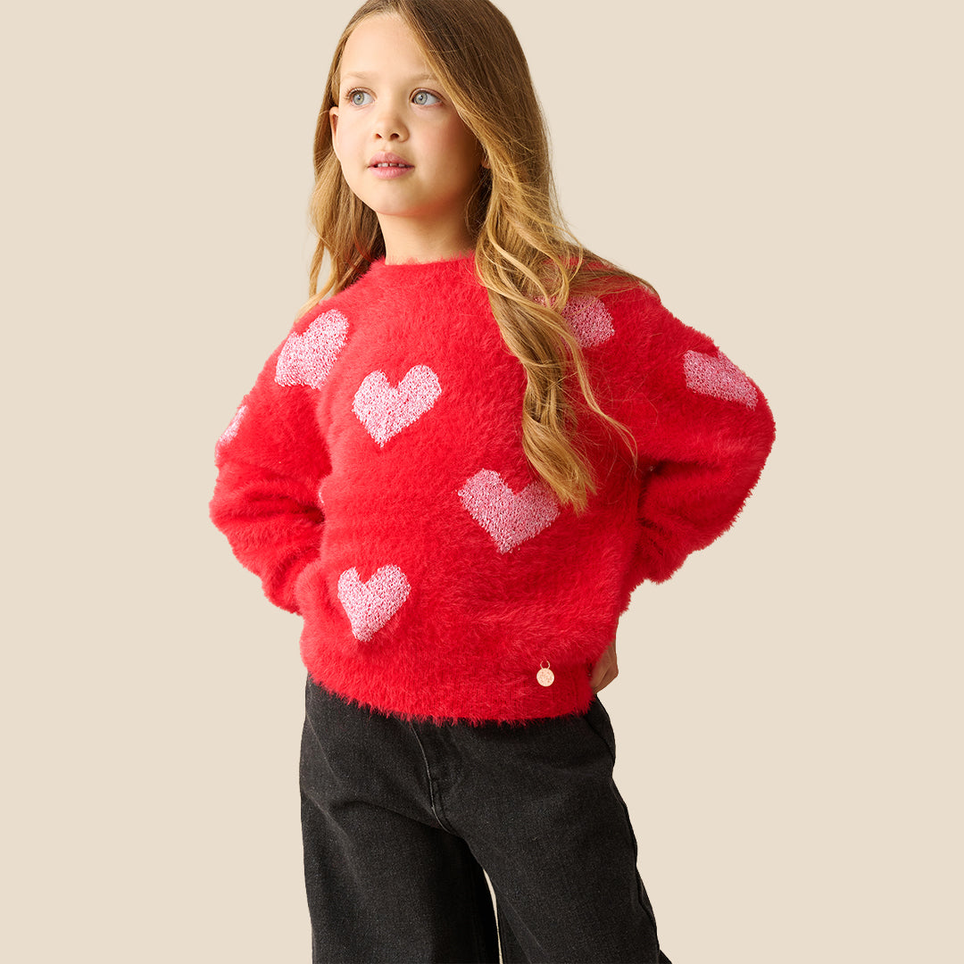 A young girl with long light brown hair wearing a fuzzy red knit sweater with light pink hearts, posing against a solid beige background.