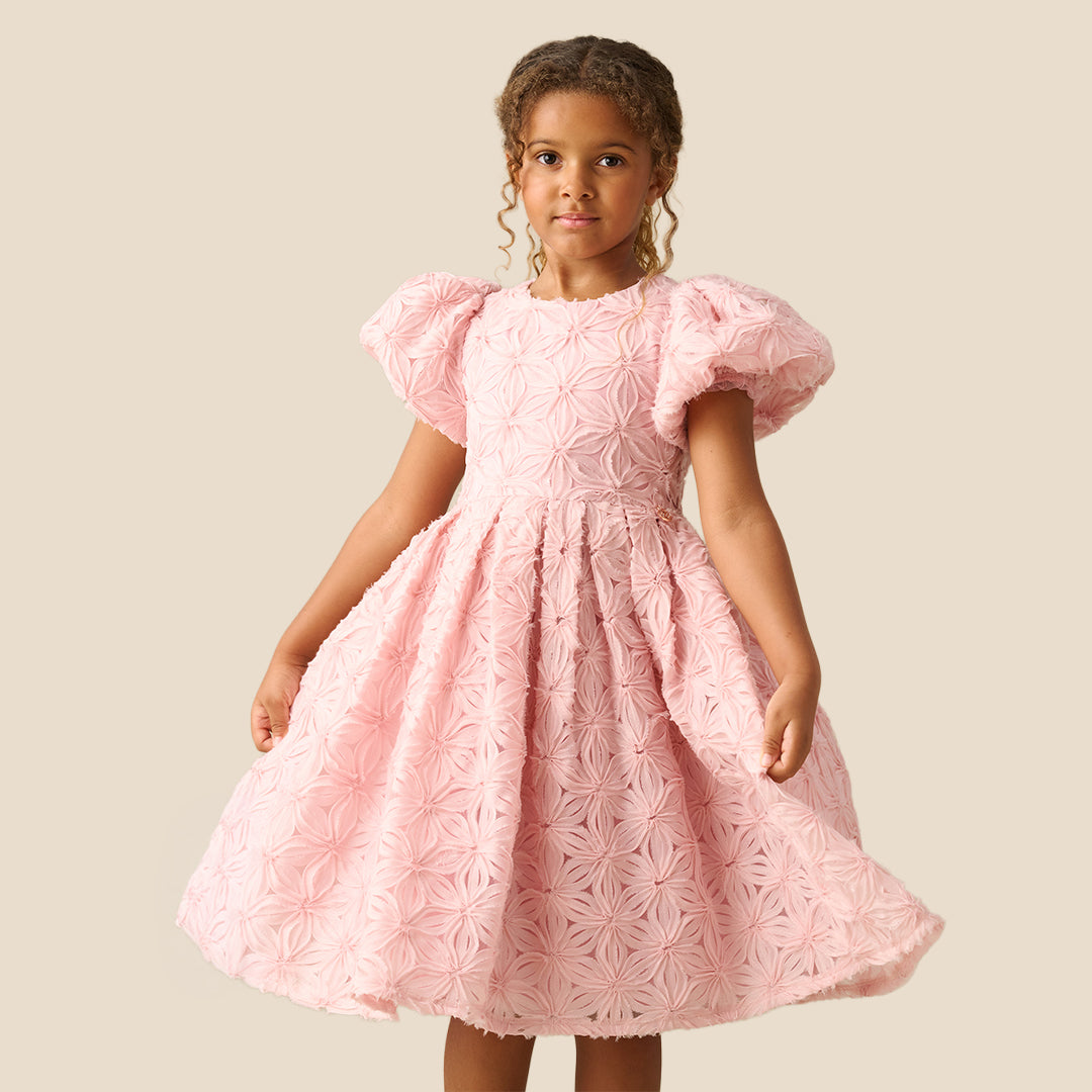 A young girl with curly brown hair wearing a light pink textured dress with puffed short sleeves and a full skirt, standing against a solid beige background.