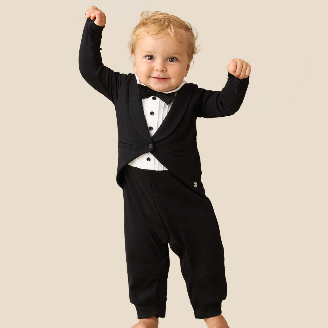 A baby wearing a black formal tuxedo-style romper with a white shirt and black bow tie, posing with arms raised against a plain beige background.