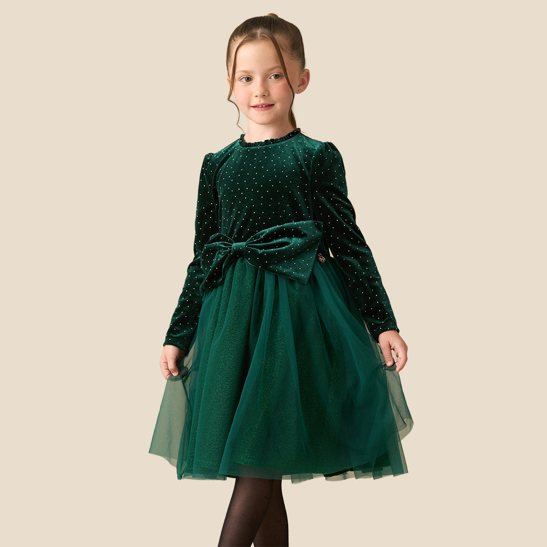 A young girl wearing a dark green velvet long-sleeve dress with a polka-dot pattern, a matching tulle skirt, and a large bow at the waist, standing against a solid beige background.