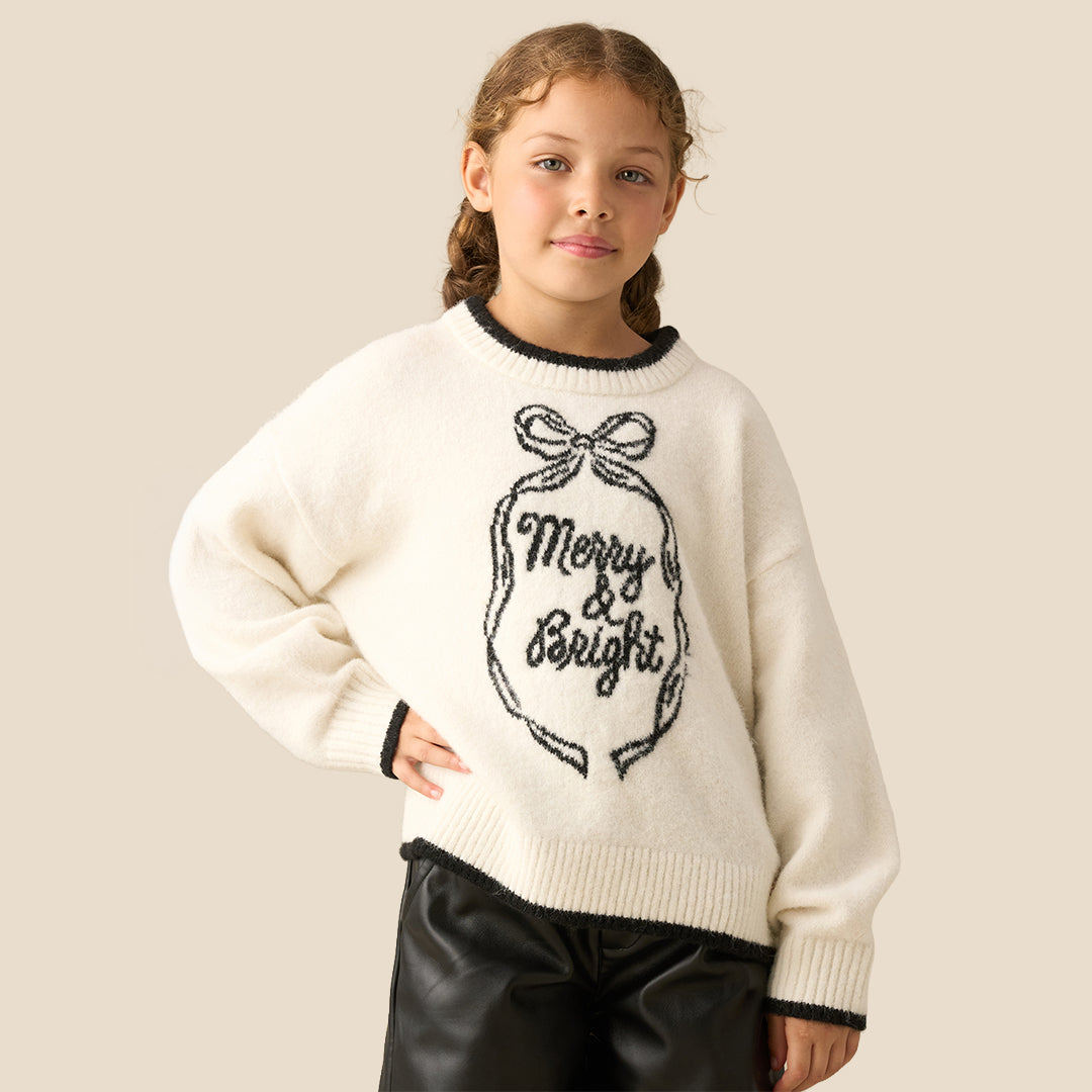 A young girl smiling while wearing a cream-colored knit sweater with a black 'Merry & Bright' ornament graphic and black trim, paired with black faux leather pants, against a solid beige background.