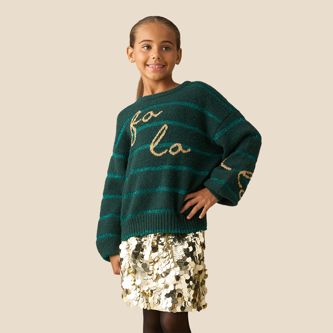 A young girl smiling while wearing a dark green knit sweater with teal horizontal stripes and gold glittery 'fo'lo' lettering on the front, paired with a gold sequined skirt, against a solid beige background.