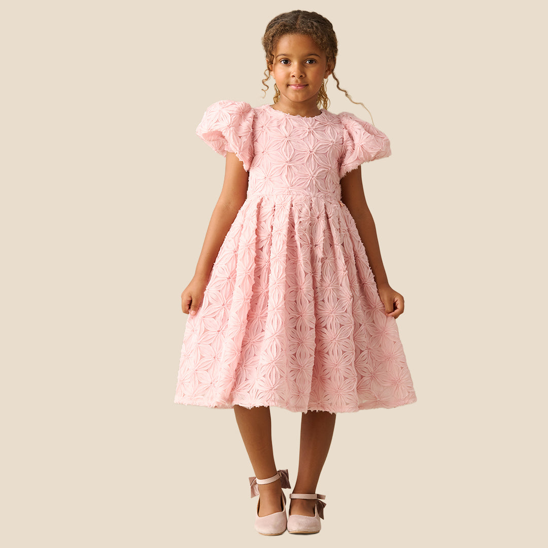 A young girl with curly brown hair wearing a light pink textured dress with puffed short sleeves and a full skirt, standing against a solid beige background.