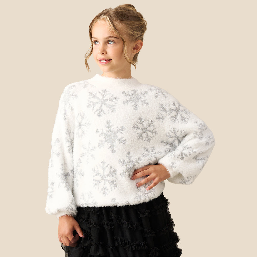A young girl with blonde hair in a bun wearing a fuzzy white long-sleeve sweater with a gray snowflake pattern, paired with a black ruffled skirt, set against a solid beige background.