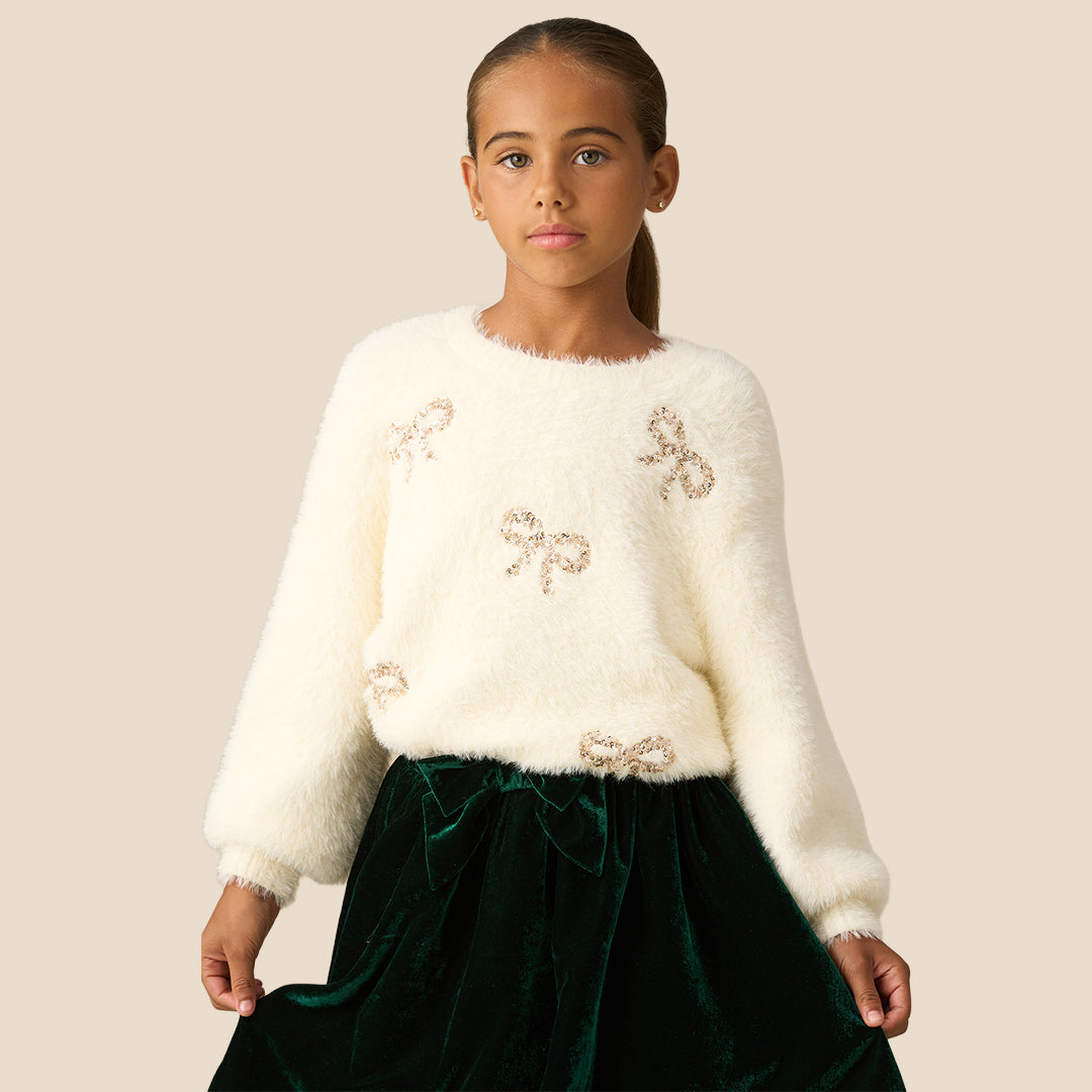 A young girl with her hair in a ponytail wearing a fuzzy off-white long-sleeve sweater with sequined bow details and a dark green velvet skirt, set against a solid beige background.