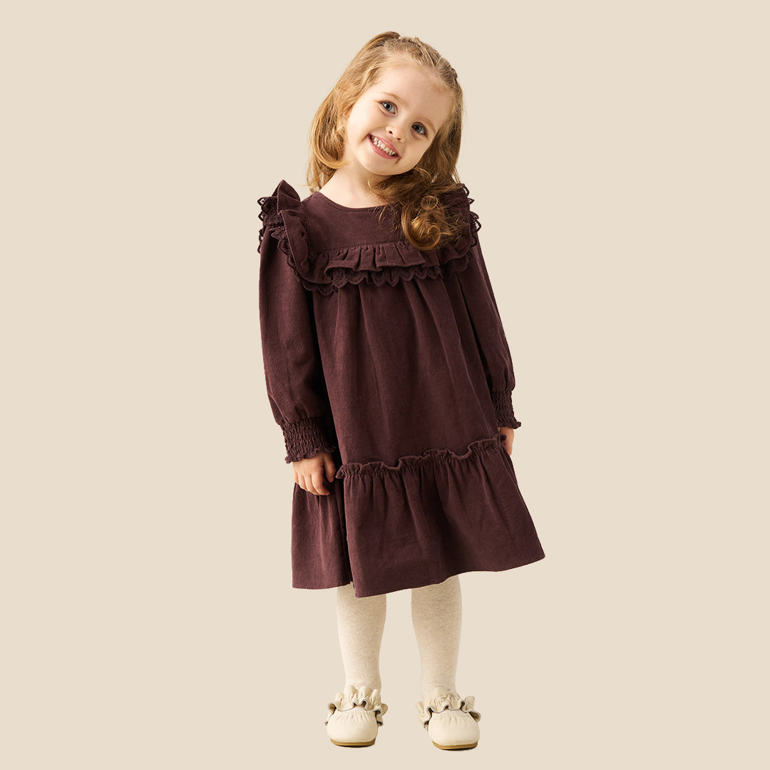 A young girl with light brown hair in pigtails stands against a solid beige background wearing a long-sleeved dark plum-brown dress with ruffles, white tights, and cream-colored shoes.