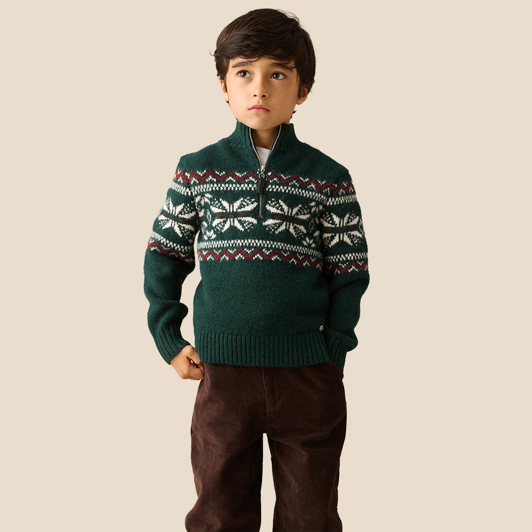 A young boy wearing a dark green knit half-zip sweater with a white and burgundy Fair Isle pattern across the chest and sleeves, paired with dark brown pants, set against a solid beige background.