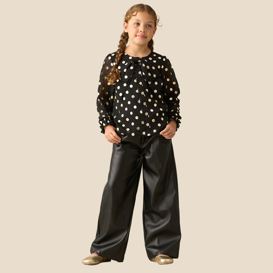 A young girl with braided hair wearing a black long-sleeve blouse with gold polka dots and black faux-leather pants, standing against a solid beige background.