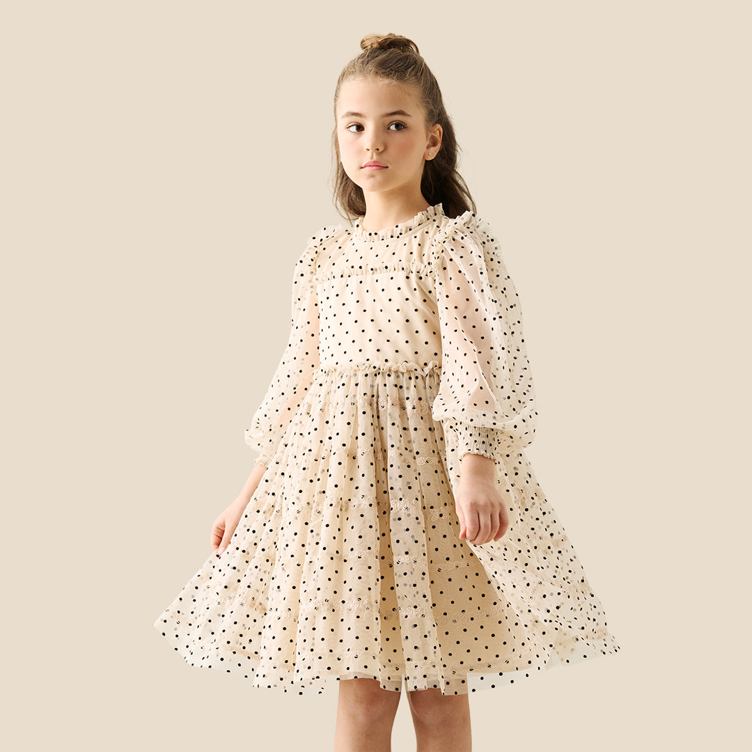 A young girl with her hair in a bun wears a cream-colored, long-sleeved tiered dress with black polka dots, standing against a solid beige background.