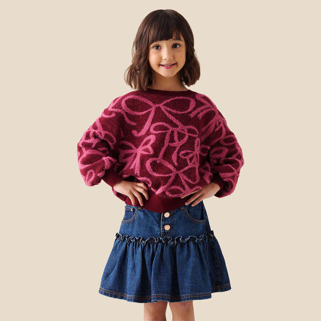 A young girl with blonde hair in a ponytail wearing a fuzzy dark red long-sleeve sweater with a pink bow pattern, paired with a blue denim skirt, against a solid beige background.