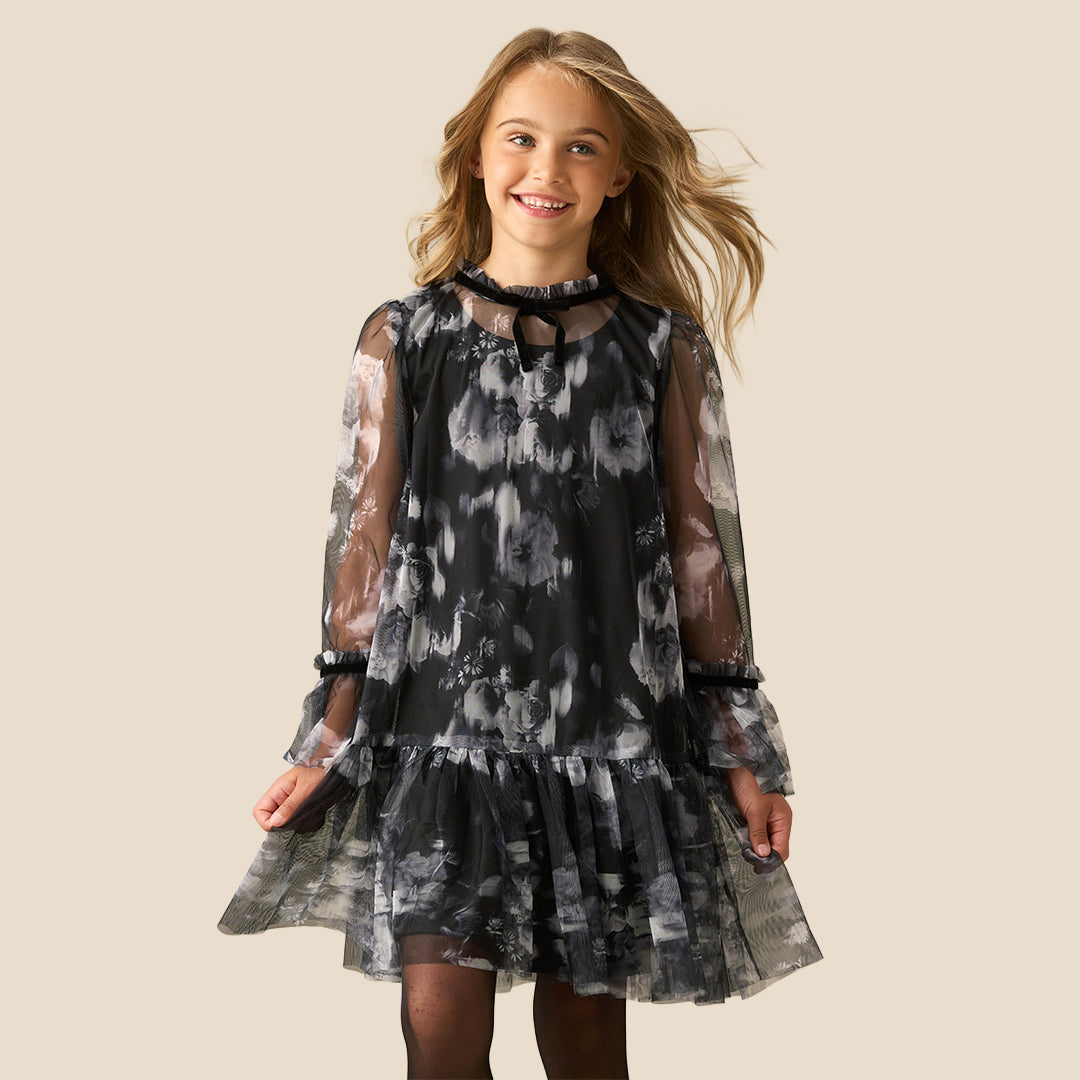 A young girl with long light brown hair is wearing a black and gray tie-dye dress with sheer long sleeves and a ruffled skirt, standing against a solid beige background.