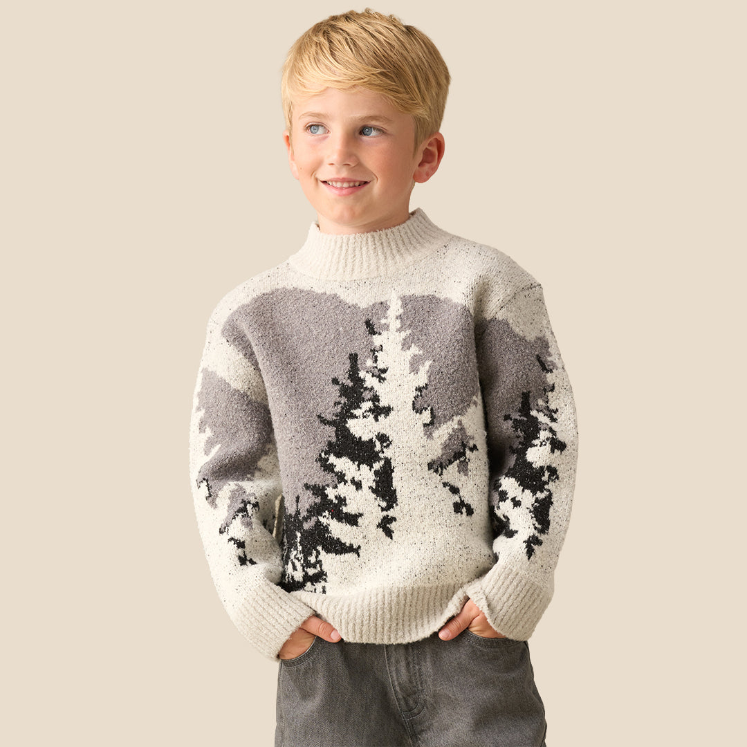 A young boy with blonde hair wearing a knit mock-neck sweater with a gray and black tree pattern, standing against a solid beige background.
