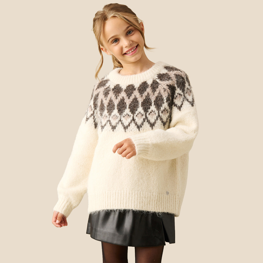 A young girl smiling while wearing a cream-colored knit sweater with a dark brown and gray geometric pattern across the chest, paired with a black skirt, against a solid beige background.