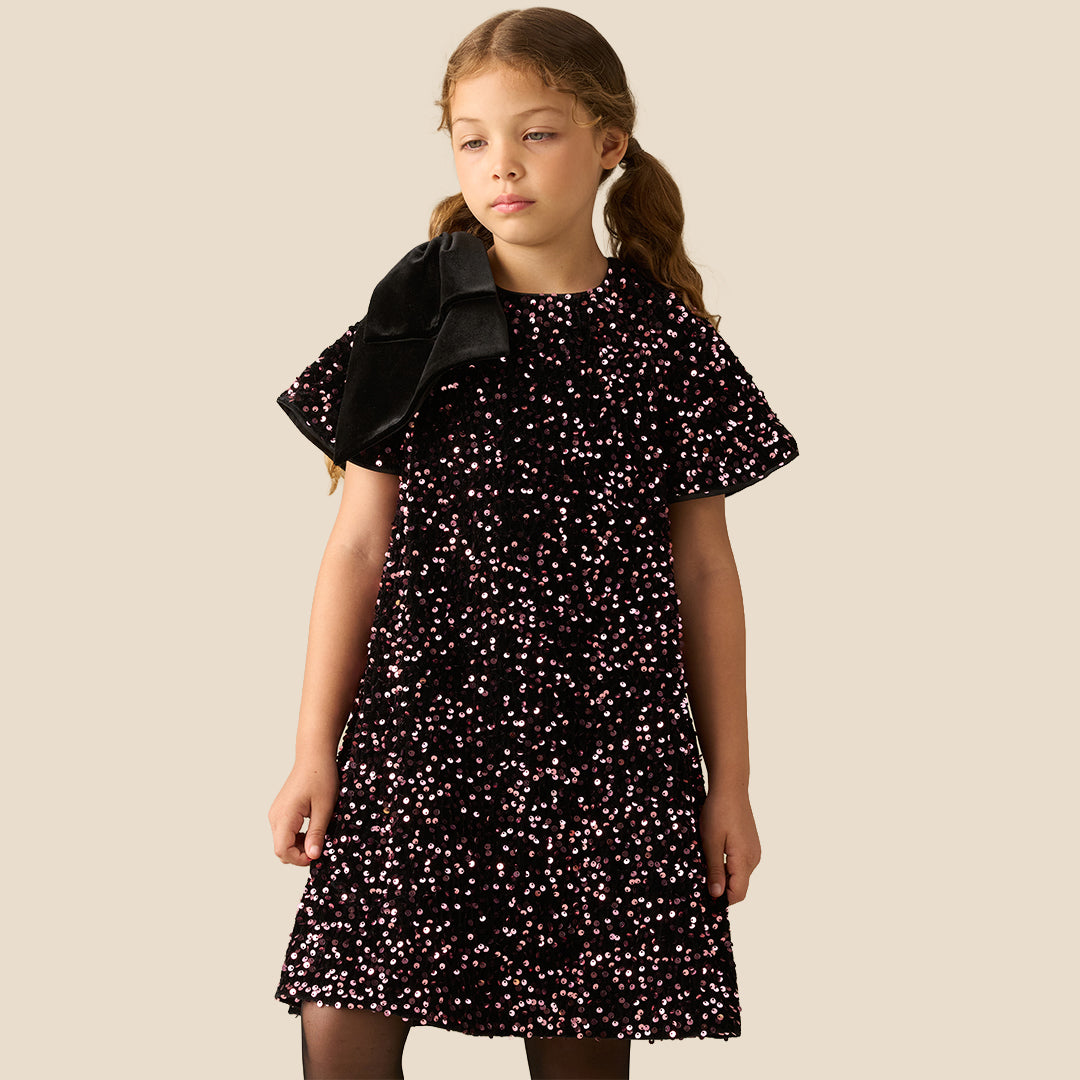 A young girl with light brown hair in pigtails stands against a solid beige background wearing a short-sleeved black dress covered in small pink and white floral patterns, with a black bow on the shoulder.