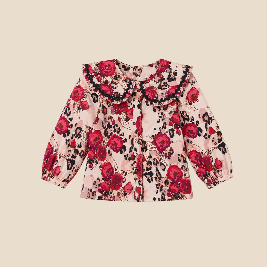 A young girl with blonde pigtails wearing a pink long-sleeve blouse with a red rose and black leopard print pattern, paired with dark pants, standing against a solid beige background.