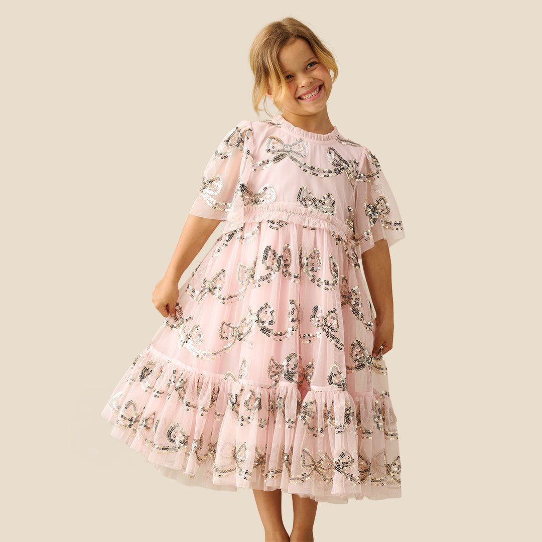 A young girl smiling and wearing a light pink tiered tulle dress with a delicate black and white floral print, set against a solid beige background.