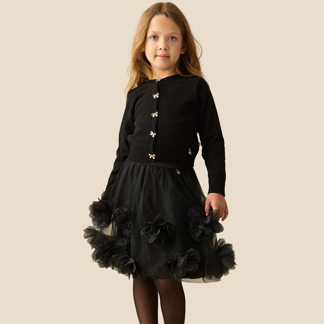 A young girl with long light brown hair wearing a black knit cardigan with gold bow-shaped buttons and a black tulle skirt, standing against a solid beige background.