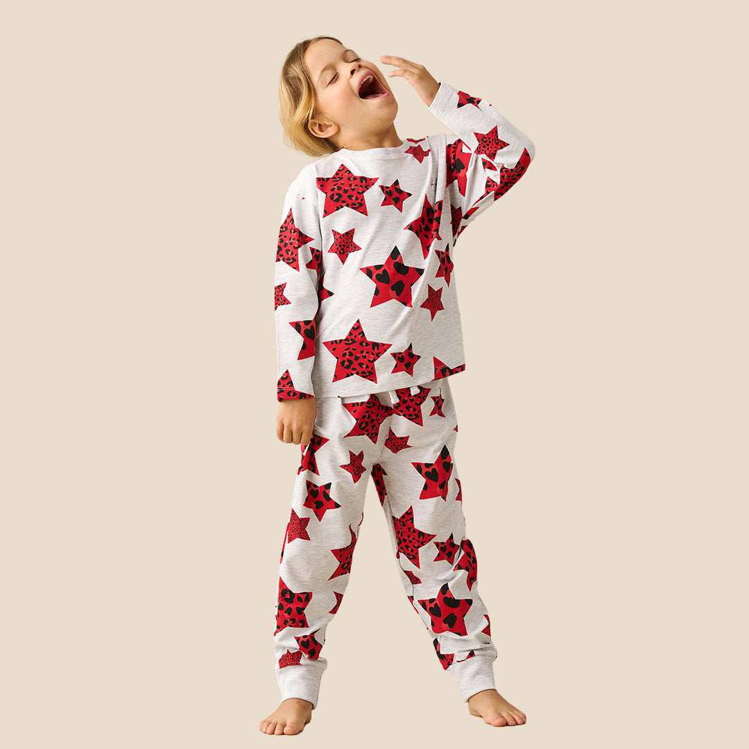 A young girl modeling a matching two-piece pajama set with a white base and a red star and ladybug print, set against a solid beige background.