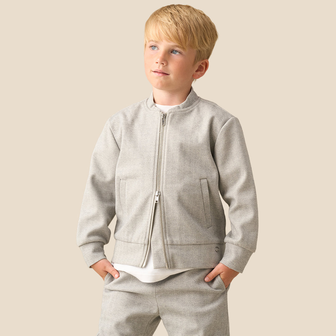 A young boy with blond hair wearing a light gray zip-up fleece jacket and matching gray sweatpants, standing against a solid beige background.