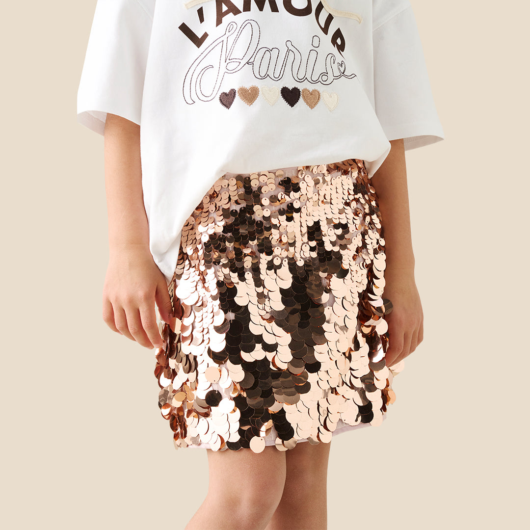 A child wearing a white graphic T-shirt and a short skirt covered in large, reversible sequins that shift between rose gold and bronze, photographed against a plain beige studio background.