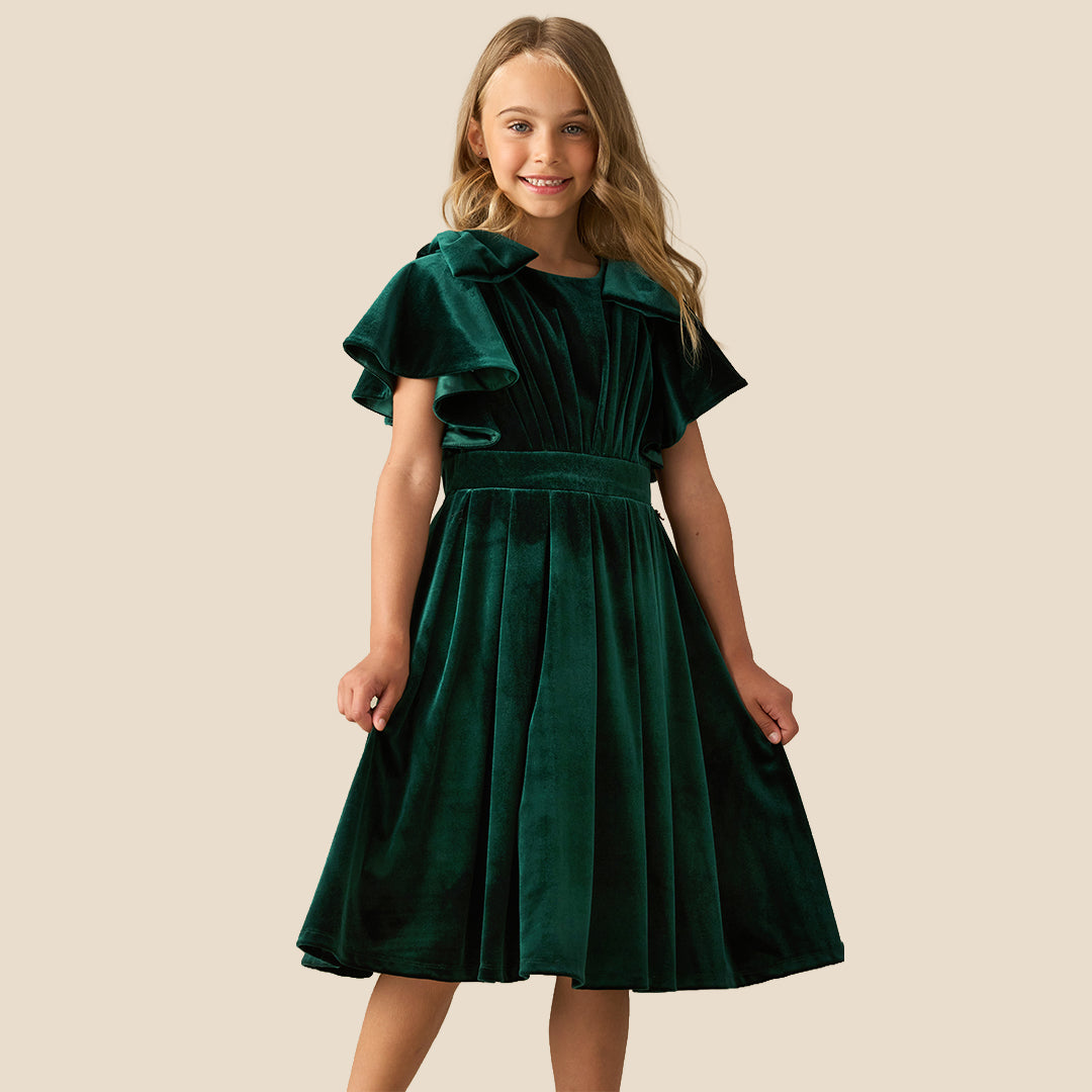 A young girl wearing a dark green velvet dress with ruffled short sleeves, a matching waist belt, and a pleated skirt, standing against a solid beige background.