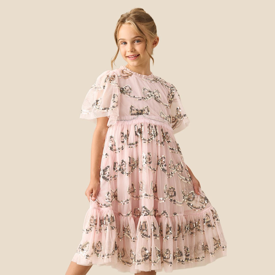 A young girl smiling and wearing a light pink tiered tulle dress with a delicate black and white floral print, set against a solid beige background.