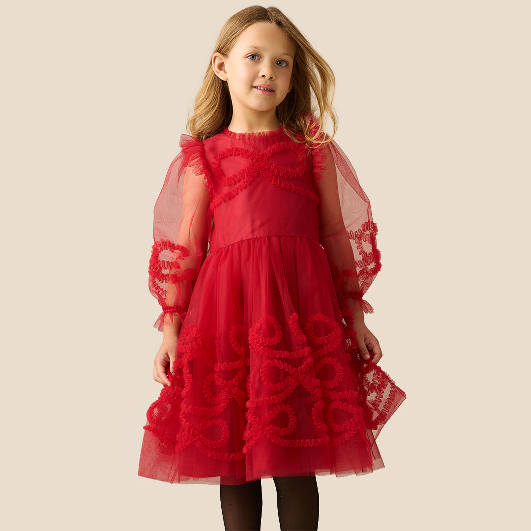 A young girl with blonde hair wearing a red tulle dress with red lace appliques and sheer flutter sleeves, standing against a solid beige background.