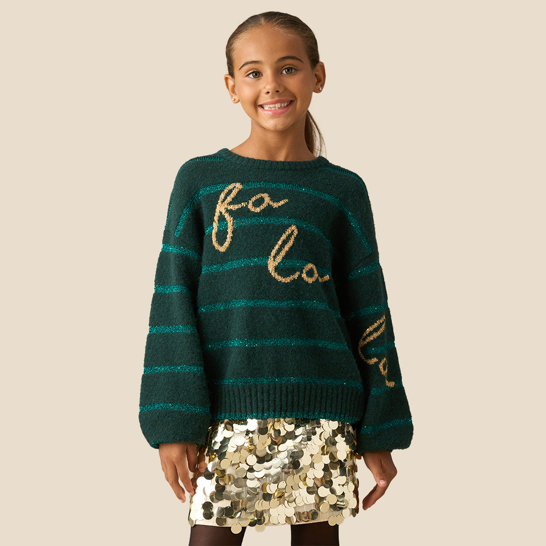 A young girl smiling while wearing a dark green knit sweater with teal horizontal stripes and gold glittery 'fo'lo' lettering on the front, paired with a gold sequined skirt, against a solid beige background.