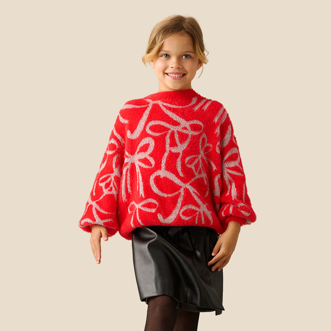 A young girl smiling while wearing a bright red fuzzy knit sweater with a white abstract pattern, paired with black bottoms, against a solid beige background.