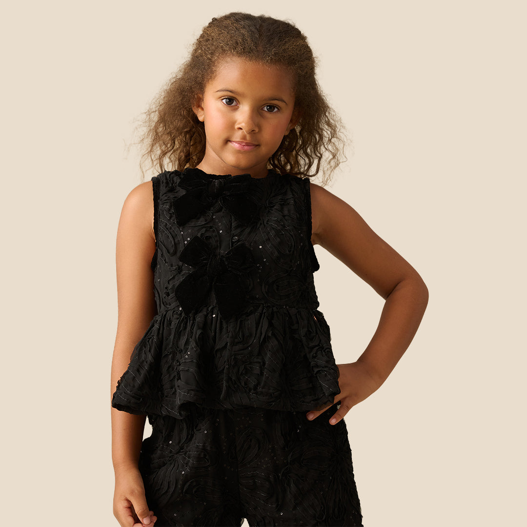 A young girl with curly brown hair wearing a sleeveless black textured dress with a ruffled peplum and bow detail, posing against a solid beige background.