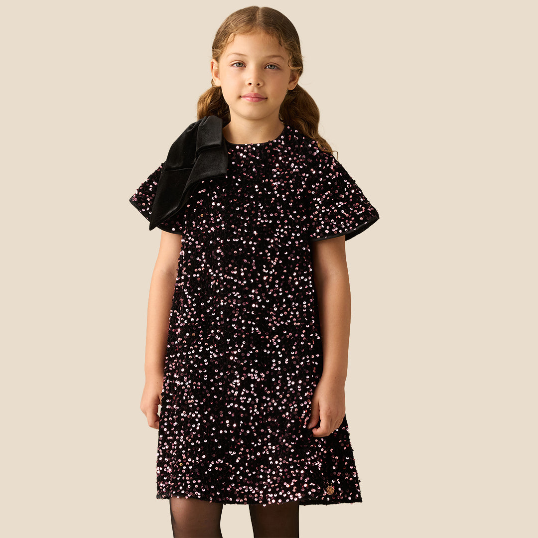 A young girl with light brown hair in pigtails stands against a solid beige background wearing a short-sleeved black dress covered in small pink and white floral patterns, with a black bow on the shoulder.