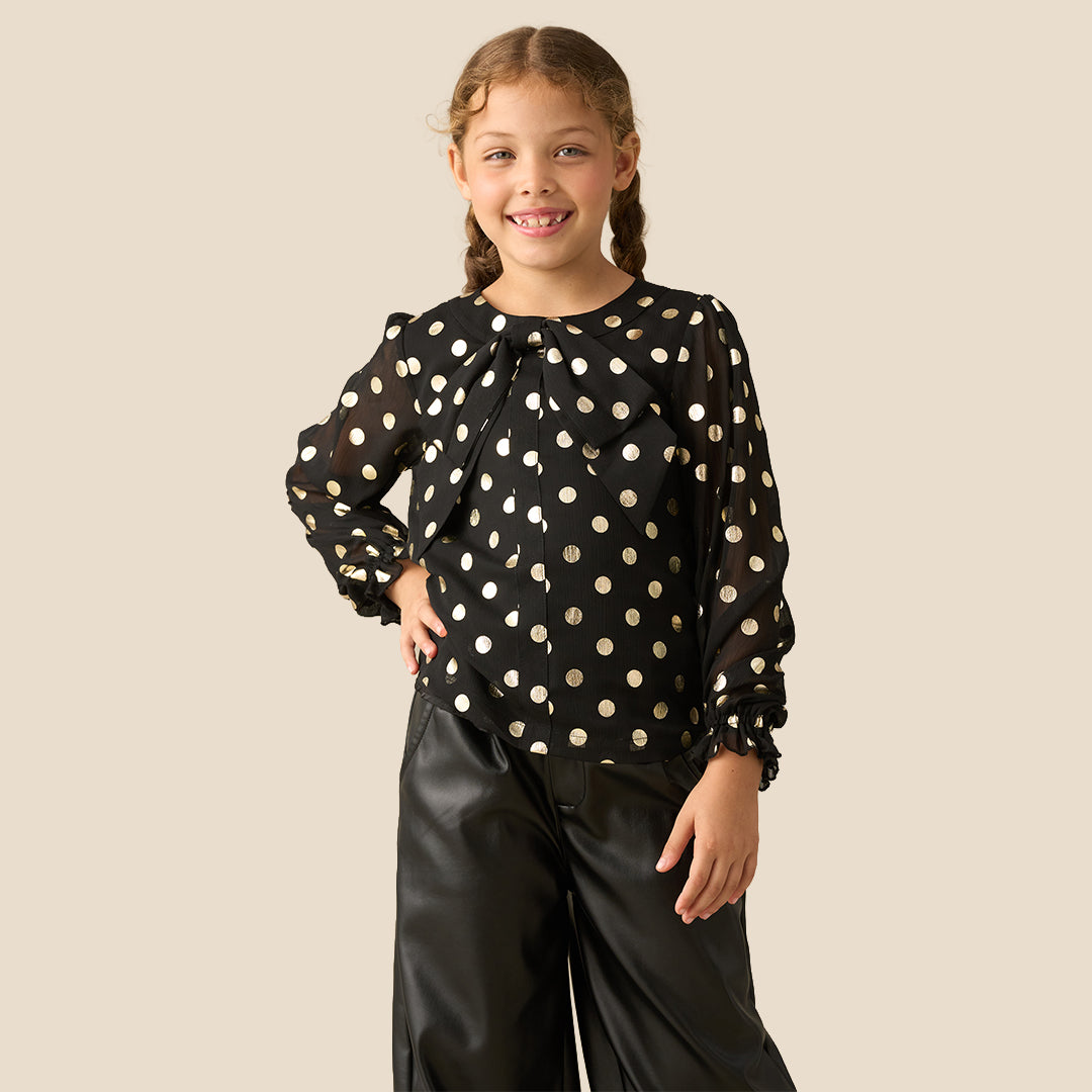 A young girl with braided hair wearing a black long-sleeve blouse with gold polka dots and black faux-leather pants, standing against a solid beige background.