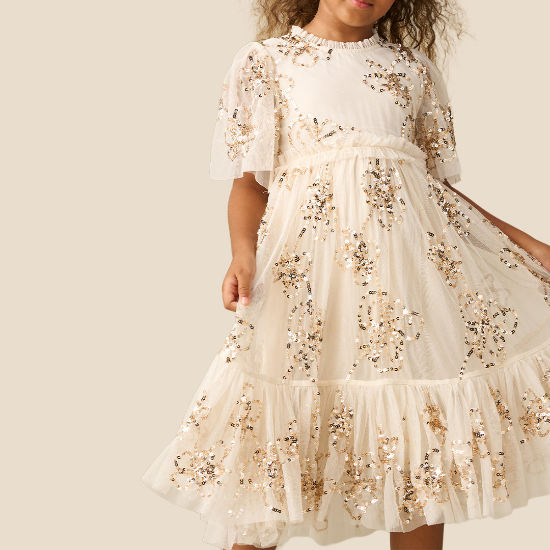 A young girl with curly brown hair wearing a tiered cream dress decorated with gold sequins and floral embroidery, standing against a solid beige background.