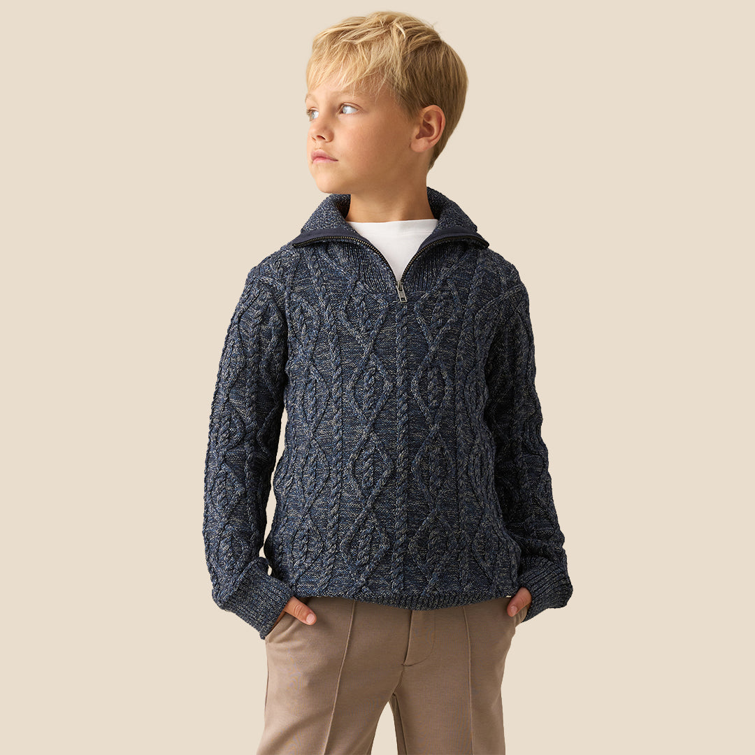 A young boy with blonde hair wearing a navy blue cable-knit mock-neck sweater and beige trousers, standing against a solid beige background.