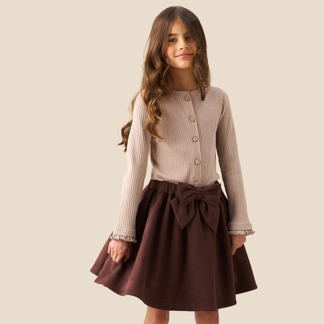 A model wearing a beige ribbed cardigan and a dark brown pleated skirt with a large bow at the waist, shown from the chest down against a solid beige background.
