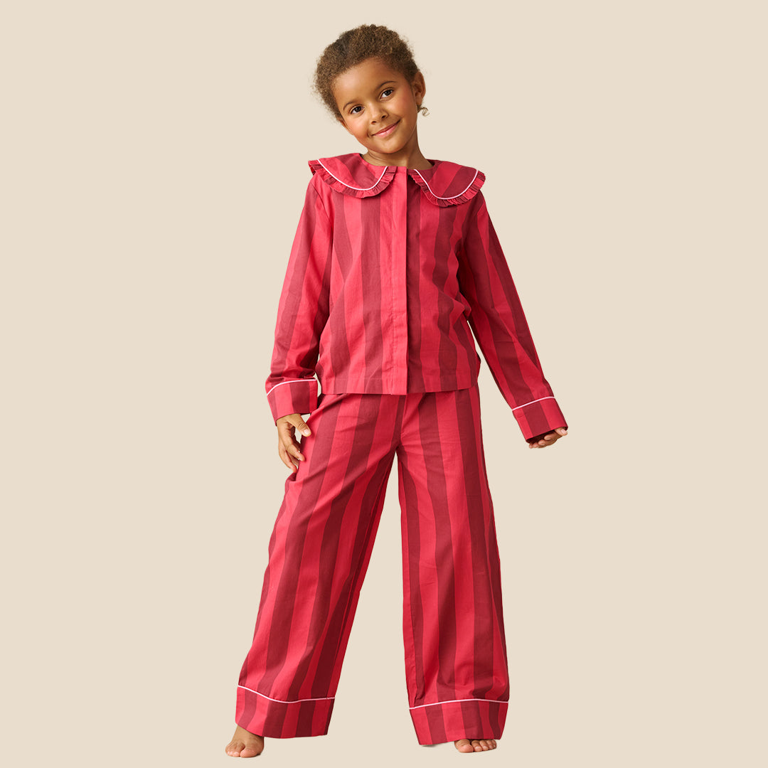 A young girl modeling a two-piece pajama set with vertical pink and red stripes, featuring a ruffled collar and white piping details, set against a solid beige background.