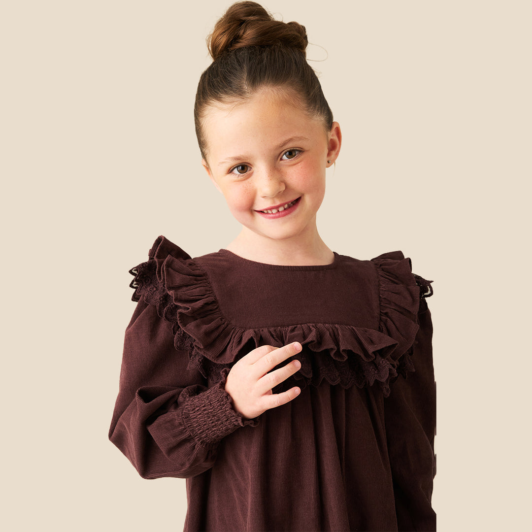 A young girl with her hair in a high bun models a dark brown long-sleeve dress with ruffled details, set against a solid light beige background.