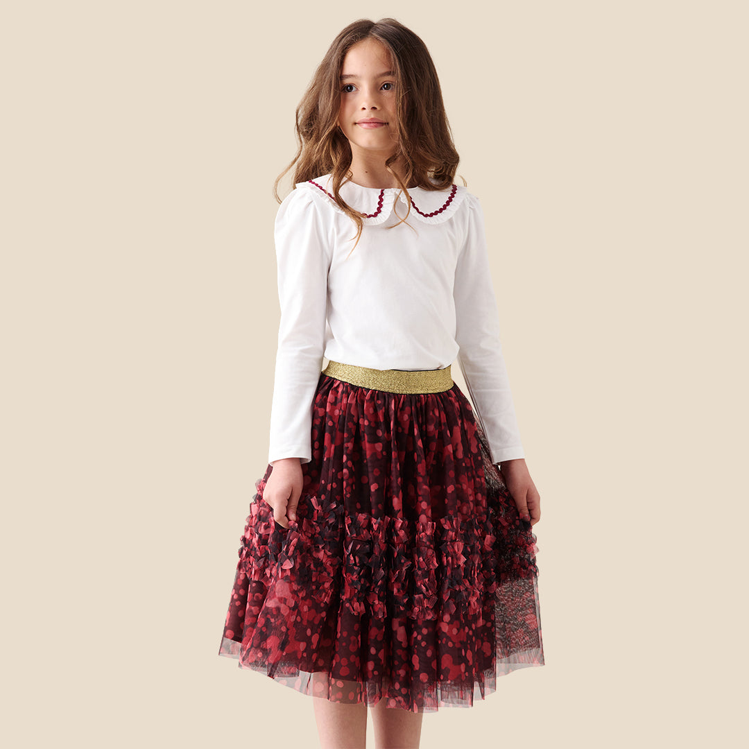 A child wearing a white long-sleeve top and a tiered tulle skirt in dark red and black floral print with a gold waistband, shown against a plain beige background.