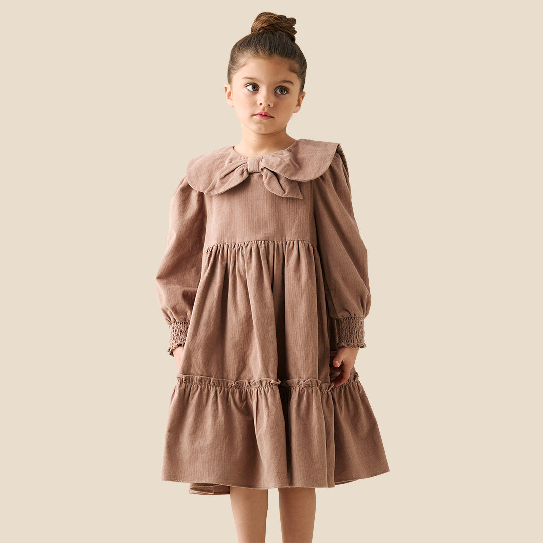 A young girl with her hair in a bun wears a brown corduroy dress with a large ruffled collar and bow, standing against a solid beige background.